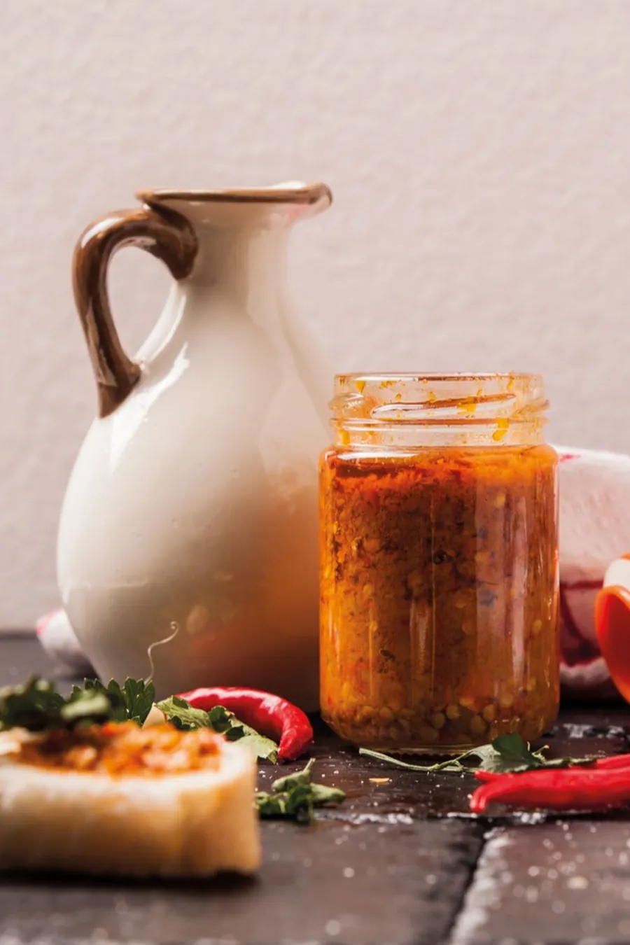 Brocca di ceramica e vasetto di conserva aperto sul tavolo, con bruschetta condita e peperoncino fresco