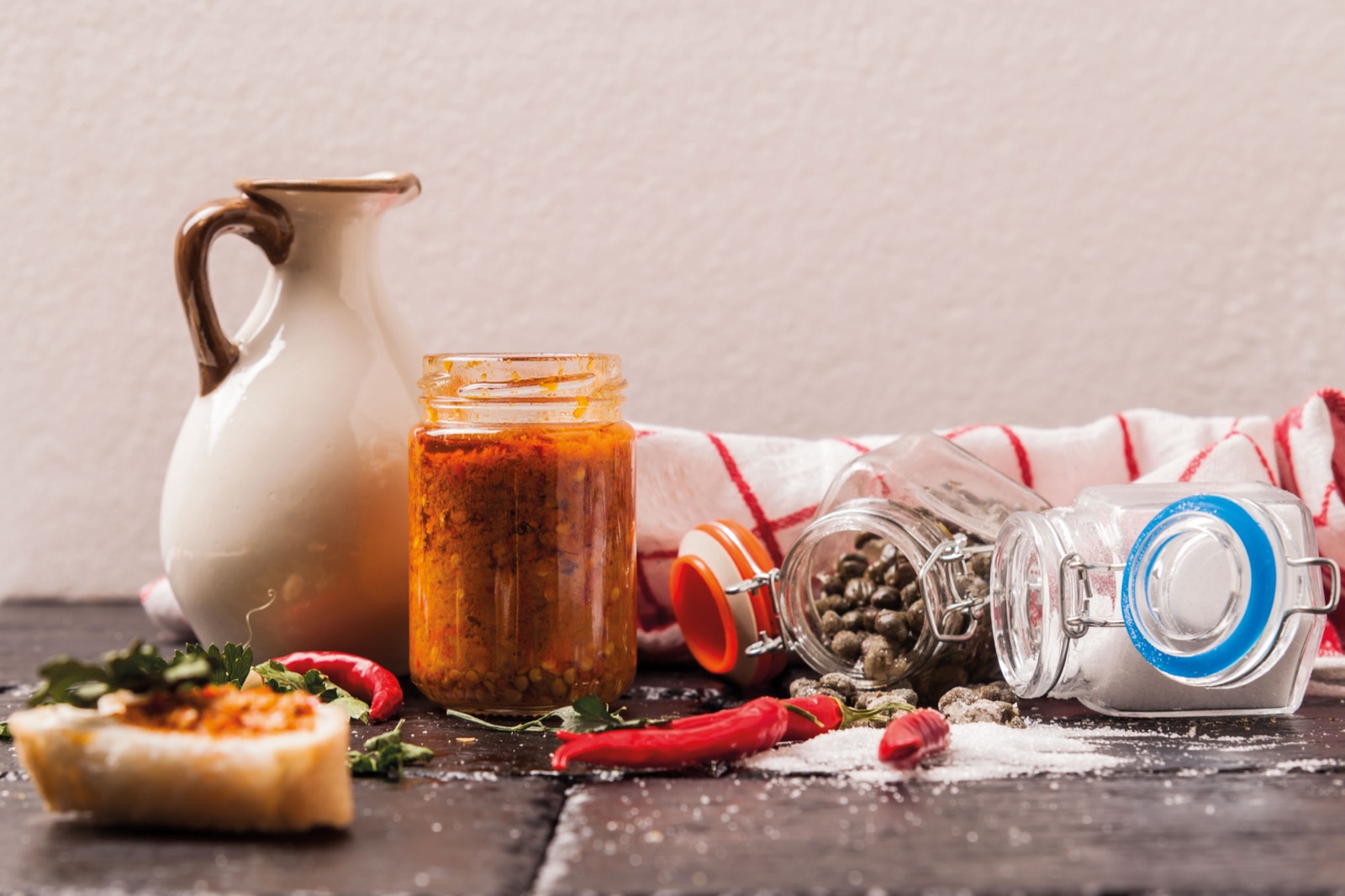 Il vasetto del Piccantino aperto sul tavolo, con peperoncino fresco, prezzemolo, capperi, sale grosso e una bruschetta condita