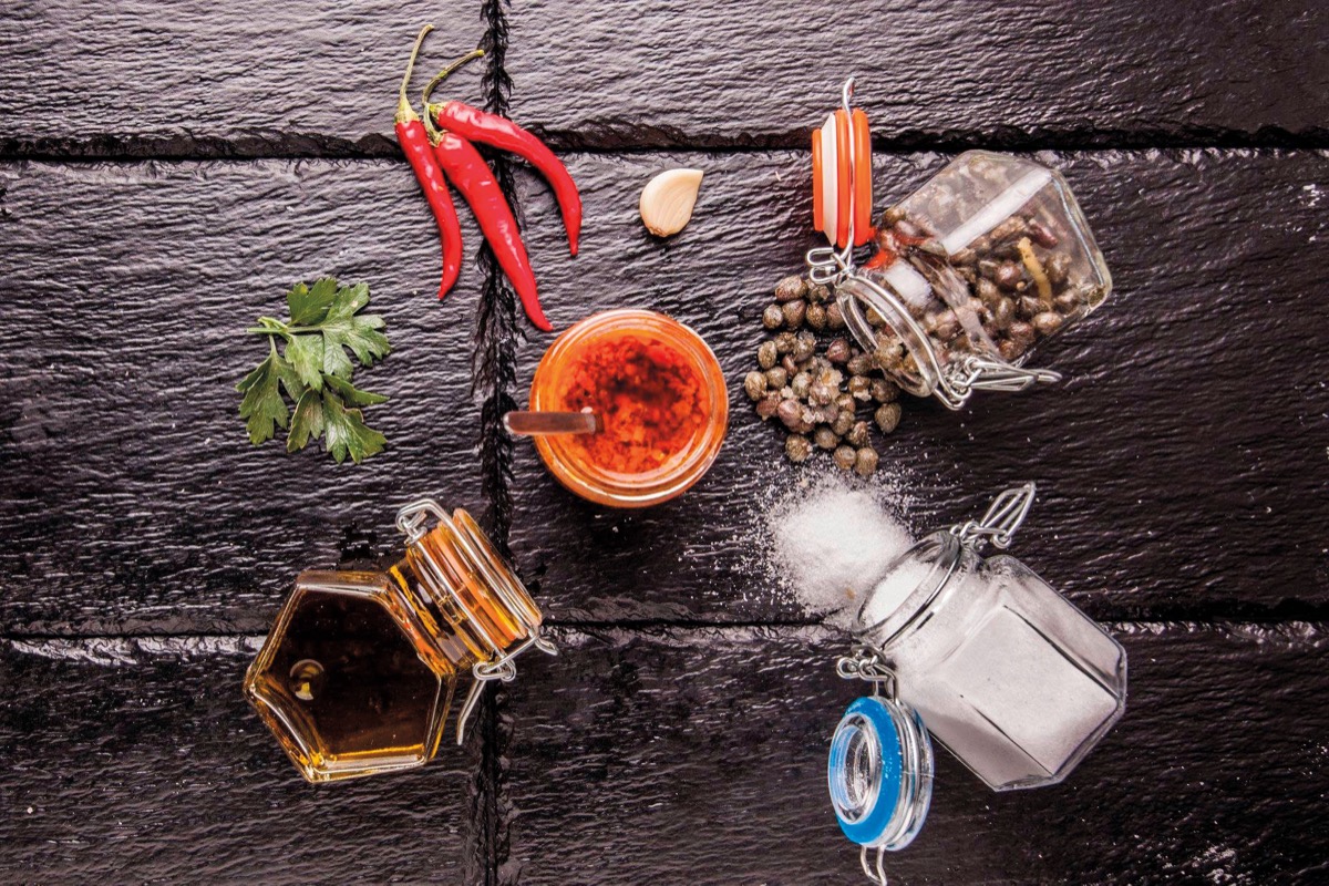 Veduta dall’alto del vasetto del Piccantino aperto con il cucchiaio, circondato da peperoncino, aglio, prezzemolo, capperi e sale